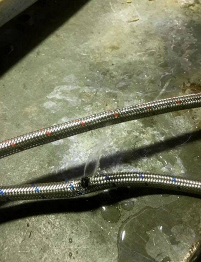 Two braided stainless-steel water supply hoses on a concrete floor, one of them burst and spraying a jet of water