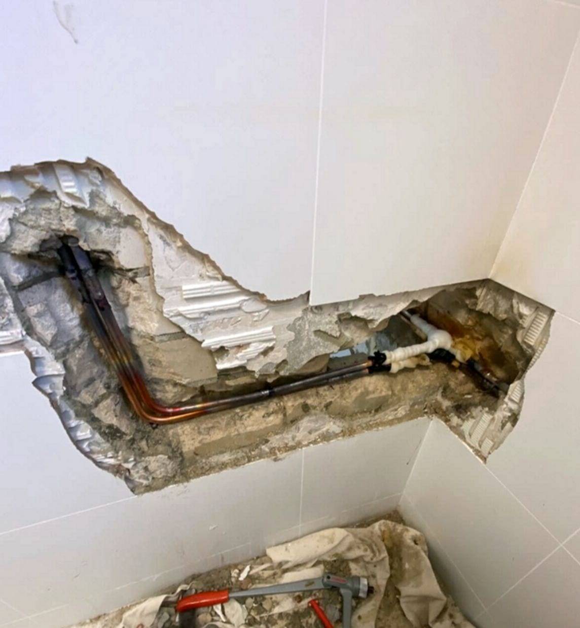 Tiled bathroom wall cut open to expose copper water lines running through a channel in the masonry, with a pipe cutter on the drop cloth below