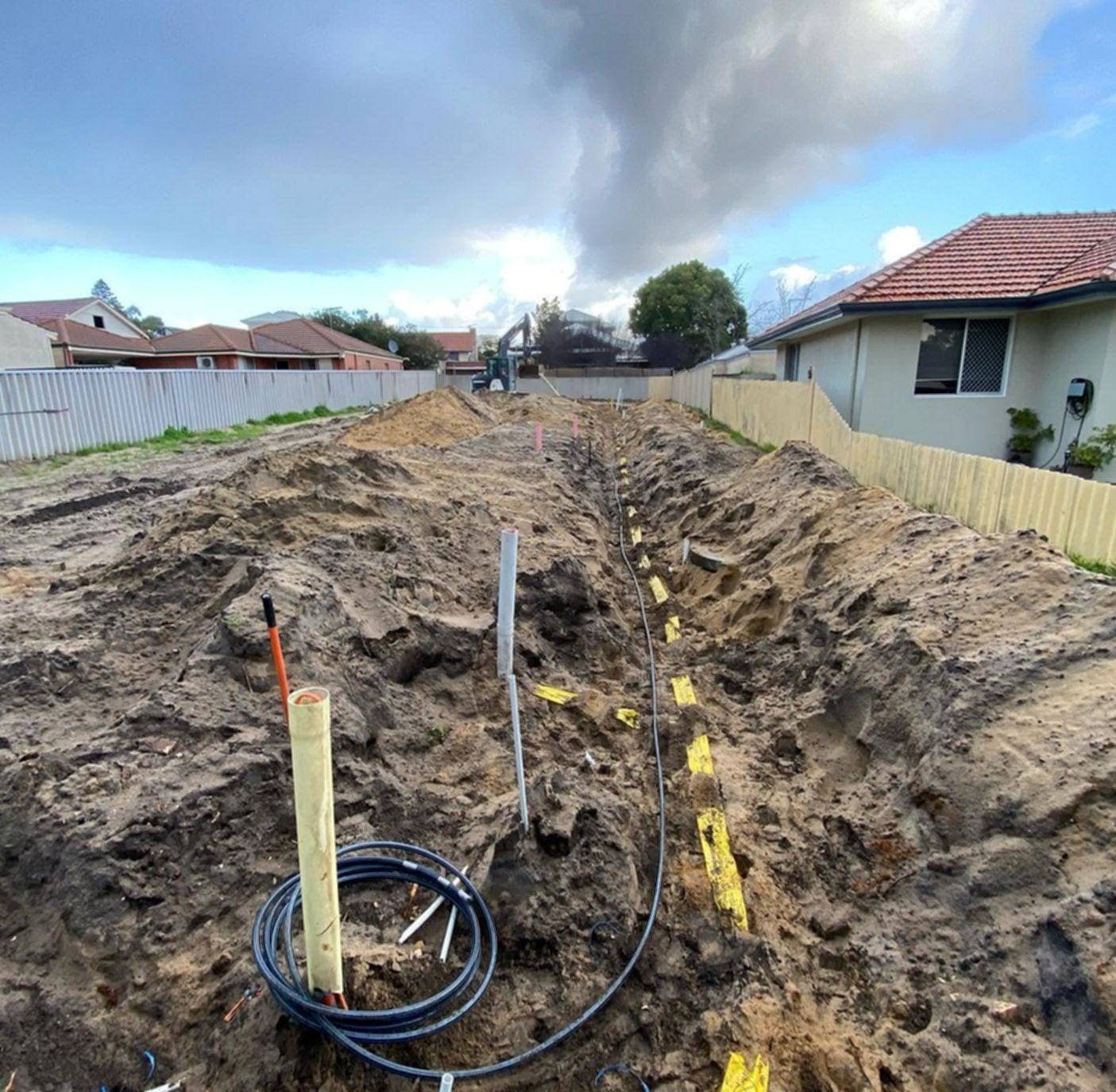 Long open trench across a cleared lot with a black water line, yellow marker tape and capped pipe risers sticking out of the soil