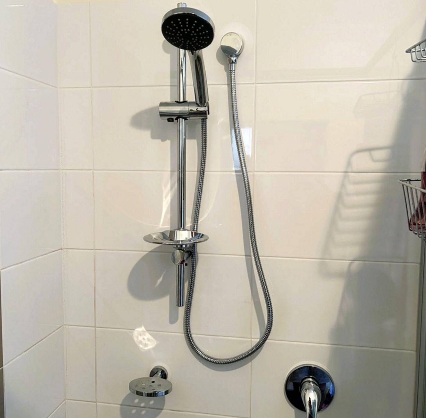 Chrome shower rail with a handheld shower head, braided hose and soap dish, with a wall-mounted mixer below, on white tile