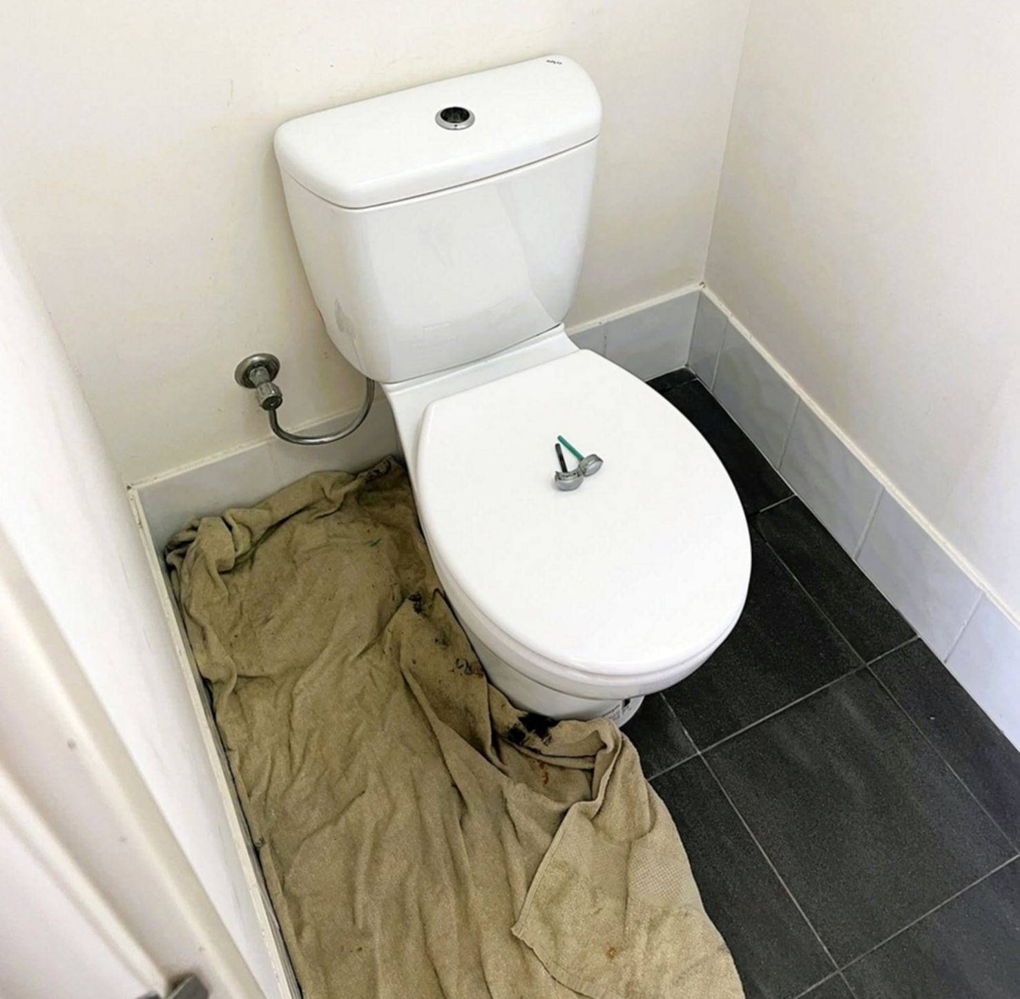 Two-piece toilet in a small bathroom with a drop cloth spread over the floor beside it and flush button parts resting on the closed lid