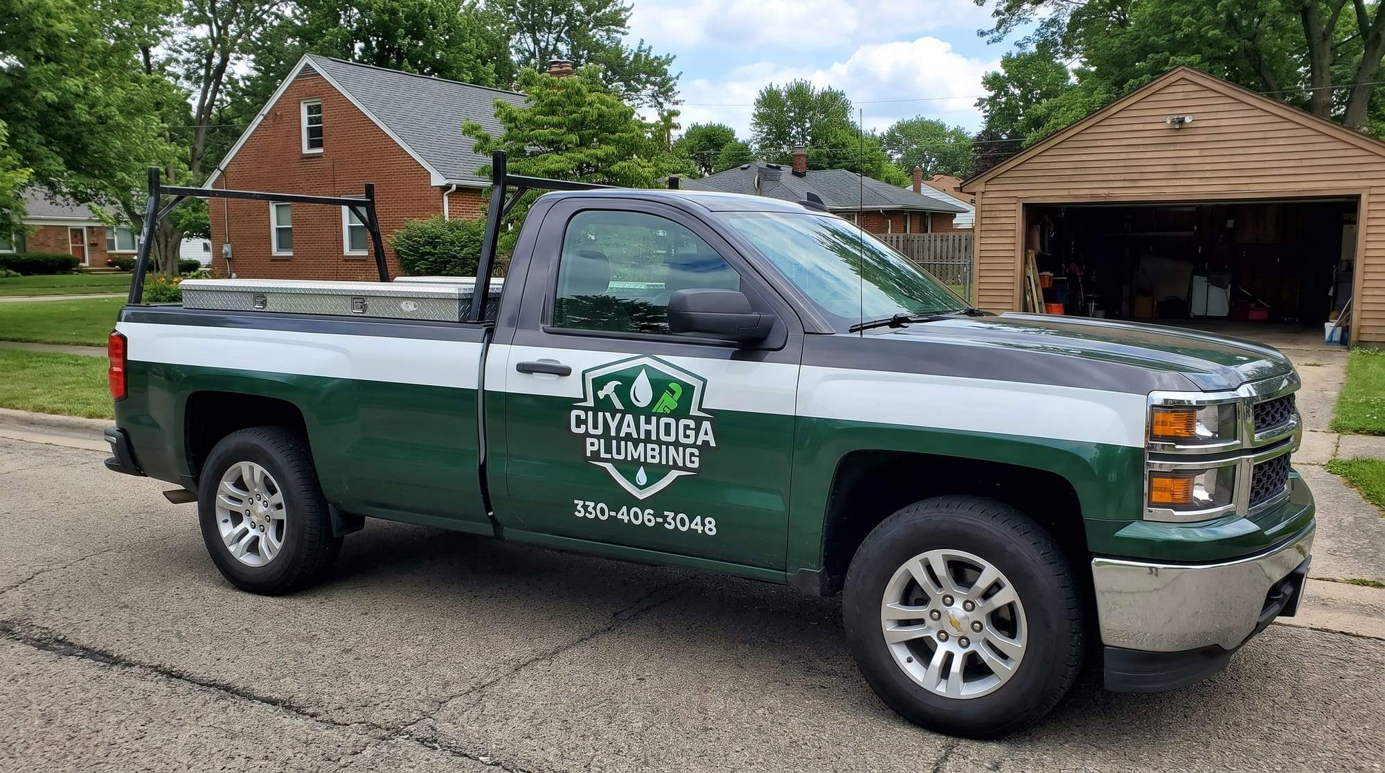 Cuyahoga Plumbing pickup truck in hunter green, white and charcoal with the company shield logo and 330-406-3048 on the door, parked on a residential street in front of a brick house